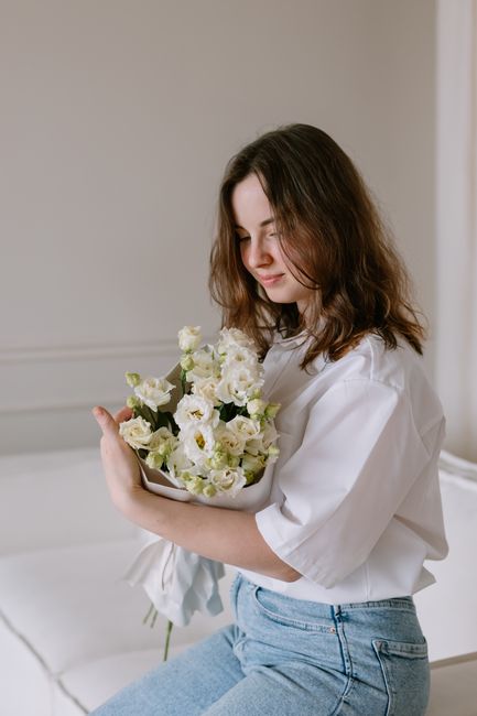 A young woman with shoulder-length brown hair, dressed in a white blouse and light blue jeans, is sitting indoors on a white surface. She is gently cradling a bouquet of fresh, white flowers with deli