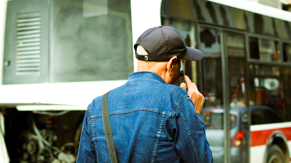 A male delivery person wearing a black cap and a denim jacket is holding a mobile phone to his ear, standing beside a large vehicle possibly used for flower deliveries. The vehicle has a reflective su