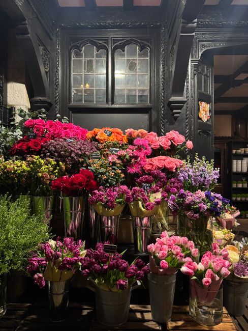 A variety of fresh flowers arranged in metal buckets and glass vases are displayed on a dark wooden shelving unit inside a florist shop. The flowers include pink, purple, red, and orange blooms such a