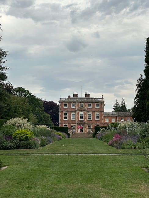 A grand red brick mansion with white-framed windows and chimneys, set amidst lush green trees and a neatly manicured lawn. The foreground features a symmetrical flower garden with vibrant, colorful bl