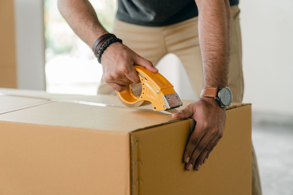 A person using a yellow utility knife to carefully cut and secure a cardboard shipping box, with their left hand holding the box steady. The individual is wearing a black wristwatch and a brown bracel